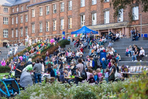 The photo shows a colourful crowd with many families with children. They are sitting on a stone staircase, doing handicrafts together at tables or chatting on the lawn.