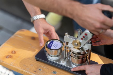 Das Foto zeigt ein Stanzgerät, mit dem ein Button gestanzt wird. Man sieht vier Hände, welche das Gerät bedienen.