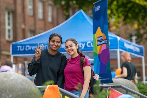 Zwei Frauen lachen in die Kamera. Im Hintergrund ist die Diversity-Flagge sowie der blaue Pavillon des Infostands des Sachgebietes Diversity Managament zu sehen.