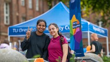 Zwei Frauen lachen in die Kamera. Im Hintergrund ist die Diversity-Flagge sowie der blaue Pavillon des Infostands des Sachgebietes Diversity Managament zu sehen.