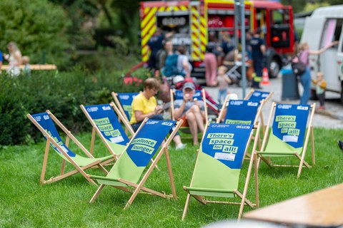Das Foto zeigt holzfarbene Liegestühle mit einer blau-grünen Sitzfläche, die auf dem Festgelände des Familienfestes stehen. Im Hintergrund sind man das Löschfahrzeug der TUD-Feuerwehr.. Auf der Sitzfläche sind verschiedene Sprüche aufgedruckt.