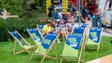 Das Foto zeigt holzfarbene Liegestühle mit einer blau-grünen Sitzfläche, die auf dem Festgelände des Familienfestes stehen. Im Hintergrund sind man das Löschfahrzeug der TUD-Feuerwehr.. Auf der Sitzfläche sind verschiedene Sprüche aufgedruckt.