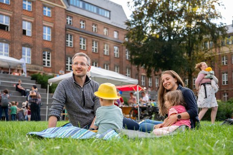 Ein Mann, eine Frau und zwei Kinder sitzen auf der Wiese auf einer Pciknickdecke.