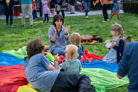 Wiese mit bunter Plane, auf der Kinder sitzen und musizieren