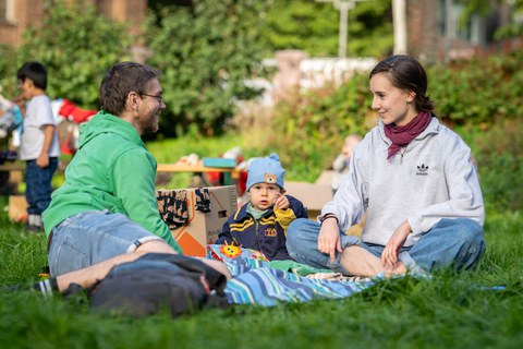 Mann und Frau mit Kleinkind in der Mitte sitzen auf einer Decke auf der Festwiese