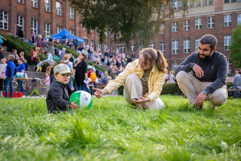 Kleiner Junge sitzt auf der Wiese und hält einen Wasserball in den Händen; Eltern sitzen daneben 