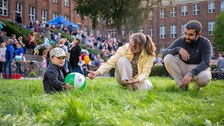 Kleiner Junge sitzt auf der Wiese und hält einen Wasserball in den Händen; Eltern sitzen daneben 