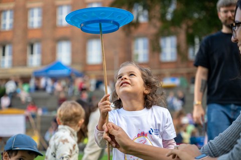 Mädchen mit weißem T-Shirt balanciert einen blauen Teller auf einem Holzstab.