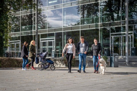 Foto mehrerer Personen vor dem HSZ der TUD. Eine Person schiebt einen Kinderwagen und eine weitere lässt sich von einem Blindenhund führen.