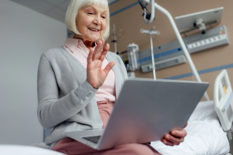 Foto einer älteren Person, die mit einem Laptop auf einem Krankenhausbett sitzt und fröhlich in den Bildschirm winkt.