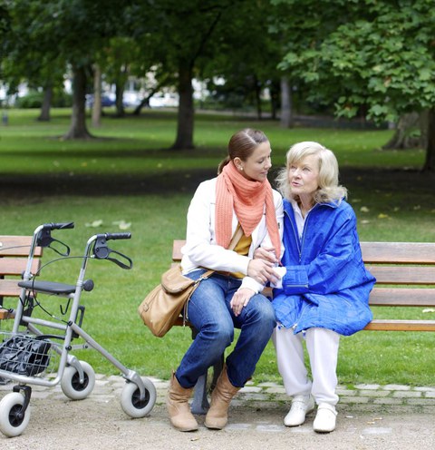 Zwei Frauen unterschiedlichen Alters sitzen auf einer Parkbank. Neben der Parkbank steht ein Rollator.