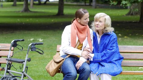 Zwei Frauen unterschiedlichen Alters sitzen auf einer Parkbank. Neben der Parkbank steht ein Rollator.