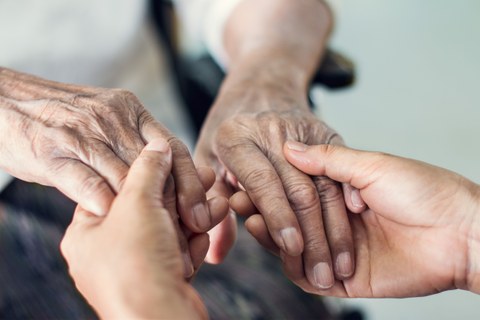 Das Foto zeigt die faltigen Hände einer älteren Person, welche in den Händen einer jüngeren Person leigen und von diesen umschlossen werden.