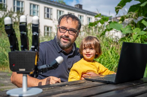 Das Foto zeigt einen Vater mit seinem Sohn. Beide sitzen an einem Tisch. Vor ihnen steht ein aufgeklappter Lptop sowie eine Roboterhand.