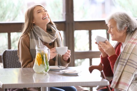Bild einer Seniorin, die mit ihrer jungen Betreuerin an einem Tisch im Café Tee trinkt.