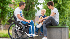 Foto zeigt einen Mann im Rollstuhl. Seine Frau und sein Kind sitzen neben ihm auf einer Bank.