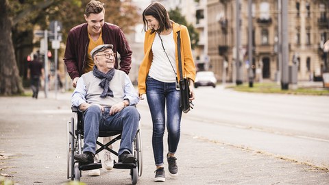 Photo shows a man and a woman walking with their wheelchair-bound grandfather.