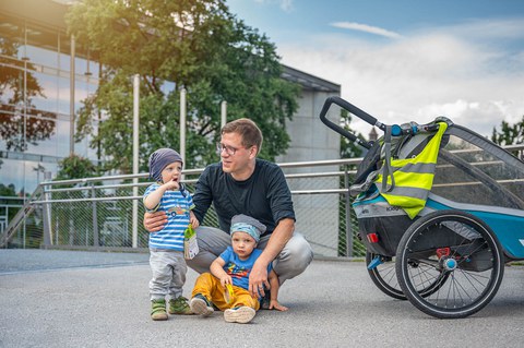 Das Foto zeigt einen Vater mit seinen beiden Kindern. Neben ihnen ist ein Kinderfahrradanhänger abgebildet.