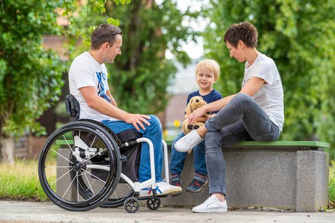 Das Foto zeigt einen jungen Mann im Rollstuhl. Daneben sitzt ein Junge. Er hält ein Stoffkuscheltier in der Hand. Ganz rechts sitzt eine Frau. Beide Erwachsenen schauen das Kind an.