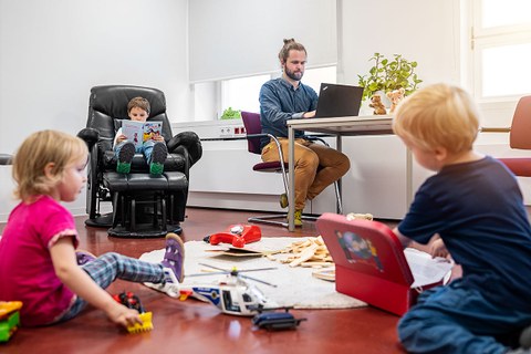 Es zeigt einen Vater, welcher im Eltern-Kind-Arbeitsraum am Laptop arbeitet und drei Kinder, die auf einem Spielteppich spielen bzw. auf einem Sessel sitzend ein Buch anschauen.
