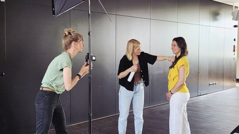 Drei Frauen vor einer schwarzen Wand. Eine Frau fotografier eine andere Frau während eine weitere der footografierten Frau die Haare richtet.