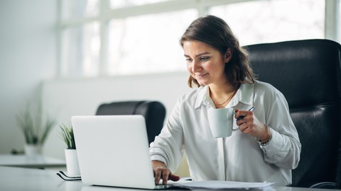 Foto einer Person im Büro am Laptop, die eine Tasse in der Hand hält.