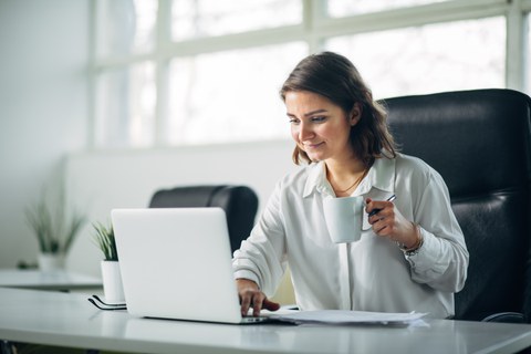 Foto einer Person im Büro am Laptop, die eine Tasse in der Hand hält.