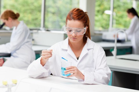 Das Foto zeigt eine Studentin mit Kittel und Schutzbrille. Sie tropft gerade mit einer Pipette blaue Flüssigkeit in ein Glas. Dadurch färbt sich die Flüssigkeit im Glas ebenfalls blau. 