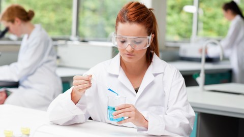 Das Foto zeigt eine Studentin mit Kittel und Schutzbrille. Sie tropft gerade mit einer Pipette blaue Flüssigkeit in ein Glas. Dadurch färbt sich die Flüssigkeit im Glas ebenfalls blau. 