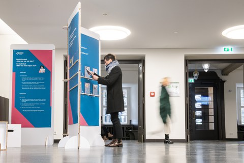 Person betrachtet Infotafeln in einer Ausstellung im Foyer, während andere vorbeigehen.