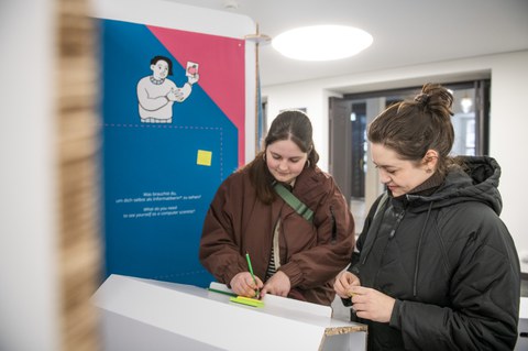 Zwei Personen schreiben an einem weißen Pult in einer Ausstellung Notizen auf farbige Zettel vor einer Infowand mit Illustration.