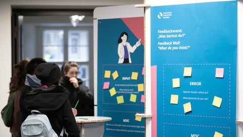 Gruppe schreibt Feedback auf bunte Zettel an einer Infowand im Foyer.