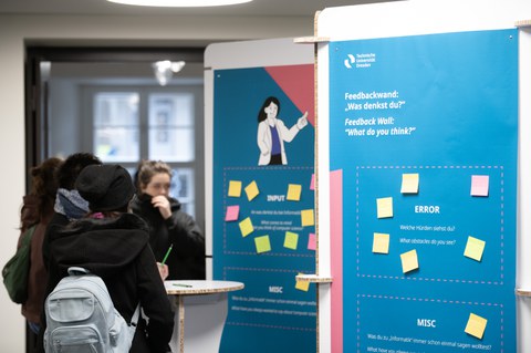 Gruppe schreibt Feedback auf bunte Zettel an einer Infowand im Foyer.