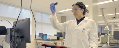 Das Bild zeigt die Wissenschaftlerin Raychelle Burks in ihrem Labor. Sie schaut auf eine Probe und trägt eine Schutzbrille und einen weißen Laborkittel. 