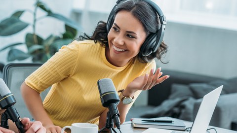 Personen mit Kopfhörern, die in einem Studio sitzen. Vor ihnen stehen Mikrofone und ein Laptop.