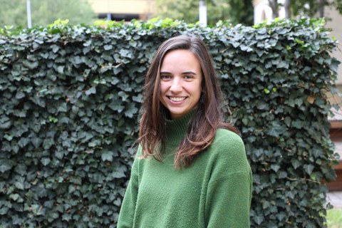 Eine lächelnde junge Frau mit langen braunen Haaren und einem grünen Pullover. Sie steht vor einer Wand mit Efeu. 