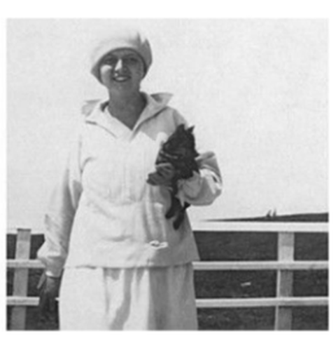 Es handelt sich um eien schwarz-weiß Fotografie. Ein Frau steht an einem Geländer. In der Hand hält sie einen schwarzen Hund. Sie trägt einen hellen Pullover und eine weiße Mütze.