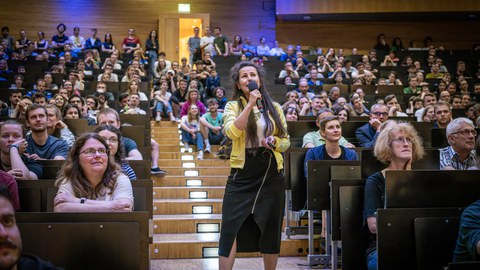 A woman is holding a microphone. She is wearing a yellow jacket and a black skirt. She is standing on a staircase in a lecture hall. To the left and right of her sits the audience.
