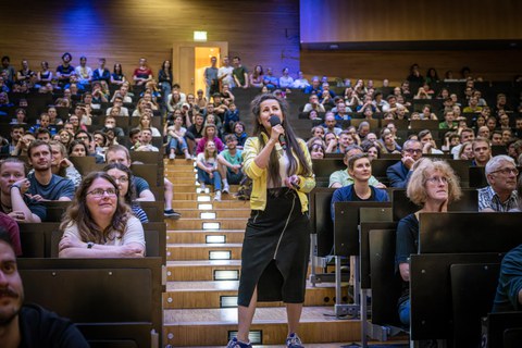 Eine Frau hält ein Mikrophon. Sie trägt eine Gelbe Jack und einen schwarzen Rock. Sie steht auf einer Treppe in einem Vortragssaal. Link und recht zur ihr sitzt das Publikum.