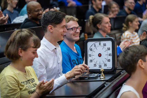 Ein Mann in einem weißen Hemd und dunklen Haaren sitzt an einem Tisch im Vorlesungssaal. Vor sich hat er eine große Stopuhr, die schwarz umrandet ist.