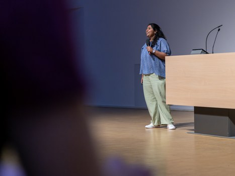 Eine Frau steht auf einer Bühne. Sie hat schwarze Haar, eine blaue Bluse, grüne Hosen und weiße Turnschuhe an. In der Hand hält sie ein Mikrophon und spricht zum Publikum.