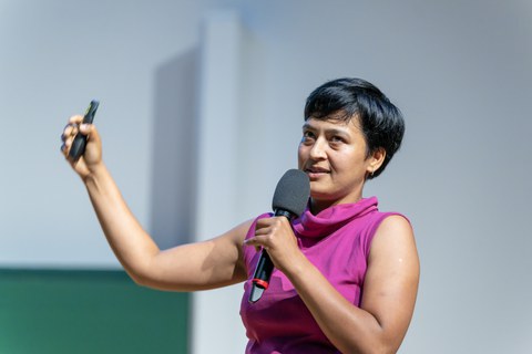 Eine Frau mit pinker kurzärmliger Blus und kurzen schwarzen Haaren. In der einen Hand hält sie ein Mikrophon, in der anderen einen Präsenter. 