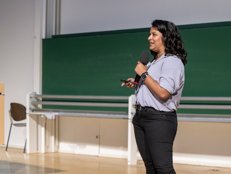 Eine Frau mit schwarzen langen Haaren, kurzärmliger hellblauer Bluse und schwarzer Hose steht auf einer Bühne. In der einen Hand hält sie ein Mikrophon in der anderen einen Präsenter