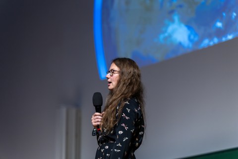 Eine Frau mit langen hellbraunen Haaren und Brille spricht in ein Mikrophon. Sie trägt ein schwarzes Kleid, welches man bis zur Hüfte sieht. Das Kleid hat weiße Applikationen.