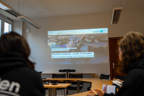 Eine Präsentation wird in einem Raum auf eine Leinwand projiziert, auf der der Titel "Verkehr studieren – an der TU Dresden. Was uns heute bewegt und morgen antreibt" zu sehen ist. Im Vordergrund sind zwei Personen angedeutet. 