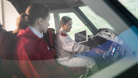 Auf dem Foto sind zwei Frauen in einer Flugzeugkabine zu sehen. Sie sprechen miteinander und sehen sich die Geräte auf dem Armaturenbrett an.