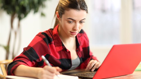 The photo shows a person sitting at a laptop and writing on a notepad.