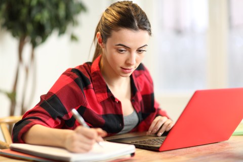 The photo shows a person sitting at a laptop and writing on a notepad.