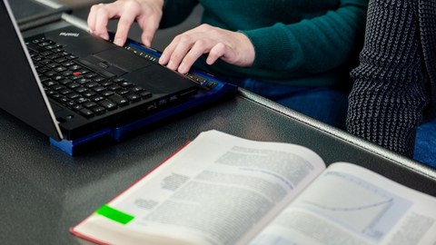 Zu sehen ist ein Laptop, an dem mit einer Braillezeile gelesen wird. Daneben ein aufgeschlagenes Buch.