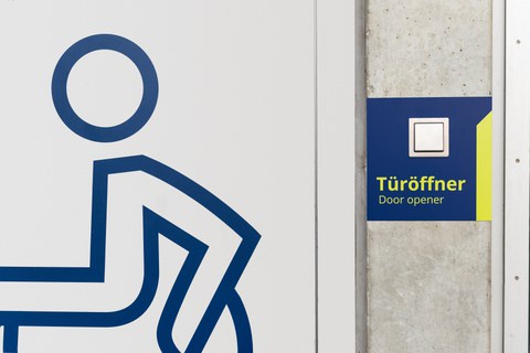  Rollstuhlsymbol auf einer Tür. Rechts davon der Schalter eines Türöffners